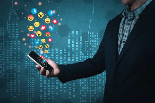 Photo of A man wearing a suit holds a mobile phone forwarded in a manner that reads a message on the screen. There are emojis emitting from the screen. Abstract background