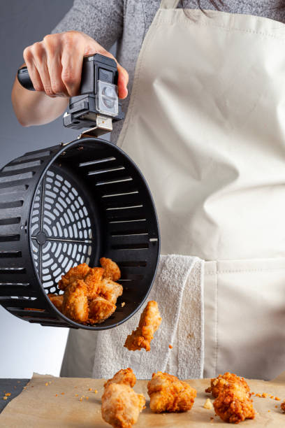 woman pouring air fried chicken nuggets on baking paper A woman is holding the handle of an air fryer oven basket with homemade fresh breaded chicken nuggets. She is pouring them onto baking paper over kitchen counter. A convenient low fat frying tool. nuggets heat stock pictures, royalty-free photos & images
