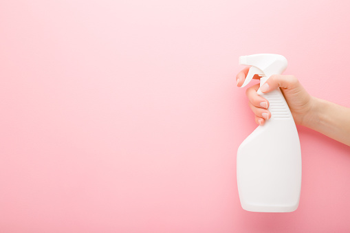 Young adult woman hand holding white spray bottle. Detergent for different surfaces in kitchen, bathroom and other rooms. Closeup. Empty place for text or logo. Light pink background. Pastel color.