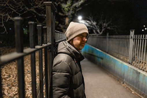 Color image depicting a homeless man in his 30s on a rundown city street at night, illuminated by street light. His expression is one of despair, sadness and depression. He is wearing a beige beanie hat and winter puffer jacket. Focus is on the man in the foreground while the background of a path and metal railing is defocused. Room for copy space.