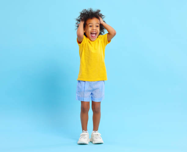 fröhliches schwarzes kind berührt haare und schreit glücklich in blauen studio - full length audio stock-fotos und bilder