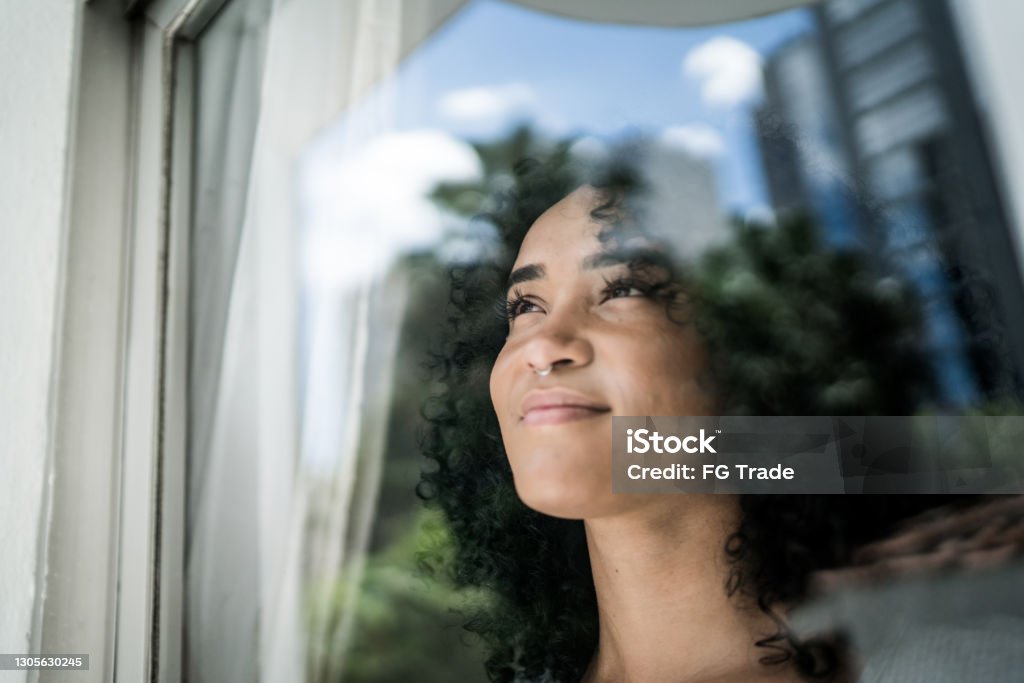 Young woman looking through window at home People Stock Photo