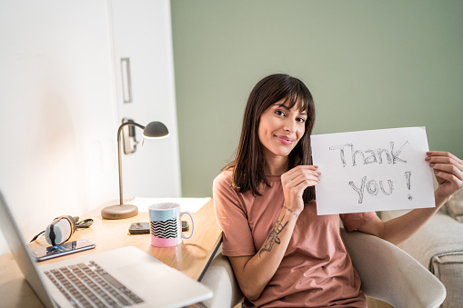 Portrait of a young woman at home holding paper with 'thank you' written