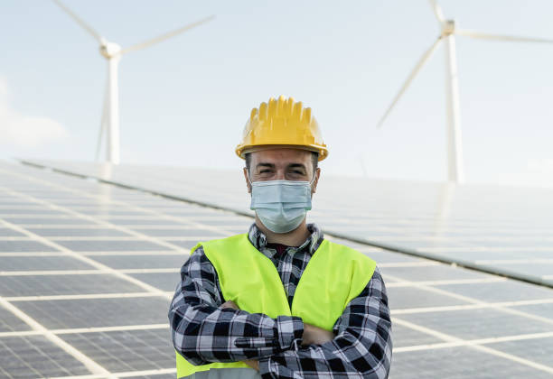 young engineer working for alternative energy with wind turbine and solar panel while wearing face mask during corona virus outbreak - innovation and green power concept - fuel and power generation wind turbine solar panel alternative energy imagens e fotografias de stock