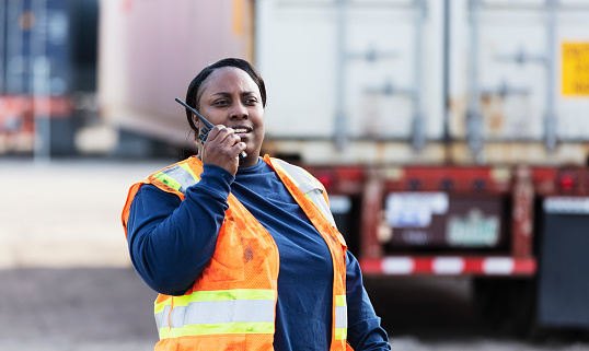 A mature African-American woman working at a freight facility, wearing a reflective vest and talking into a walkie talkie. Semi-trucks and cargo containers are out of focus in the background.