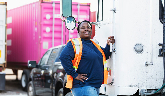 A mature African-American woman wearing a safety vest working at a commercial dock. She is a truck driver about to board her semi-truck. Cargo containers are out of focus in the background. She is looking at the camera, hand on her hip, smiling.