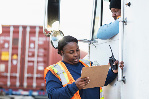 Two African-American workers in their 40s at a shipping port conversing. One is a truck driver, leaning out the open window of his semi-truck. He is talking to a woman standing next to the truck, a dock worker or manager coordinating deliveries. They are looking at the clipboard she is holding.