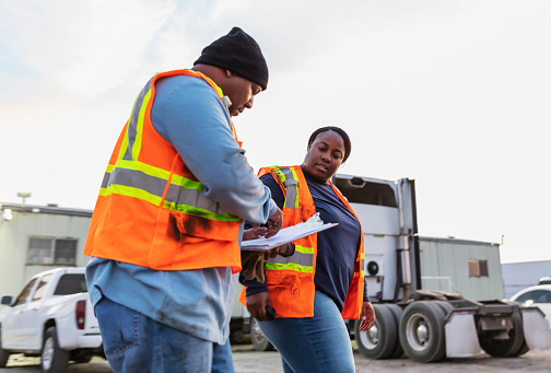 Two mature African-American workers at a freight transportation company conversing, looking at the clipboard that the man is holding. Semi-trucks and trailers with cargo containers are in the background.