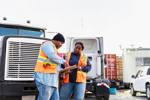 Two mature African-American workers at a freight transportation company conversing, looking at the clipboard that the man is holding. Semi-trucks and trailers with cargo containers are in the background.