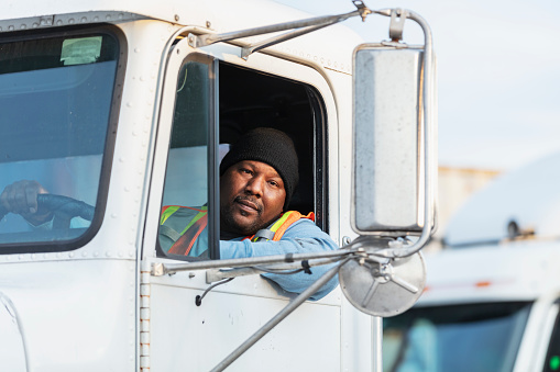 A mature African-American man in his 40s driving a semi-truck. He is sitting in the driver's seat, looking out the open window at the camera with a serious expression.