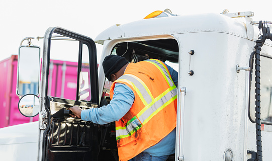 An African-American man in his 40s working as a truck driver, wearing a reflective vest, climbs into the driver's seat of his semi-truck.