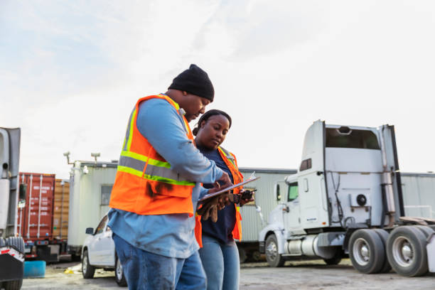 Two workers at freight company talking,, with clipboard Two mature African-American workers at a freight transportation company conversing, looking at the clipboard that the man is holding. Semi-trucks and trailers with cargo containers are in the background. trucking stock pictures, royalty-free photos & images