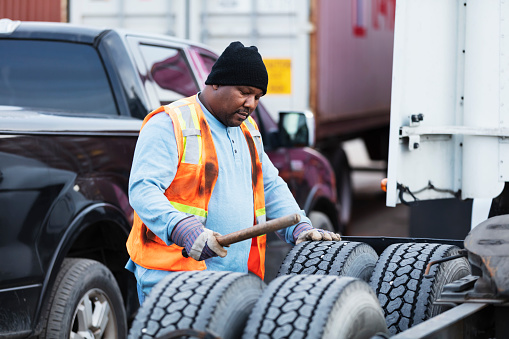 A mature African-American man in his 40s working as a truck driver, examining the tires of his semi-truck. He is using a wooden stick to make sure they have enough air pressure.