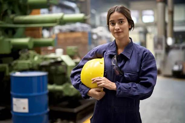 Partial front view of Caucasian woman in coveralls holding work helmet and pausing to look at camera as she walks through foundry.