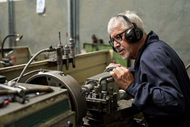 Working Portrait of Senior Male Operating Machinery Partial side view of Caucasian man in coveralls, eyeglasses, and ear protectors concentrating on production of castings in metal foundry. foundry photos stock pictures, royalty-free photos & images