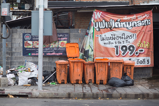 Orange garbage bins and waste on sidewalk in Bangkok Ladprao.. Sidewalk is very dirty. All wall are banners of restaurant offers. Scene is on Wanghin Road