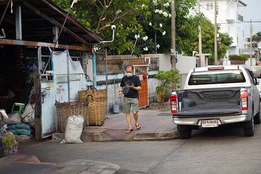 Mature thai man in casual clothings is walking while using mobile. Man is walking on sidewalk of residential area of Bangkok Ladprao between Chokchai 4 and Nakniwat. A car is parked in street. Man is wearing a protective face mask