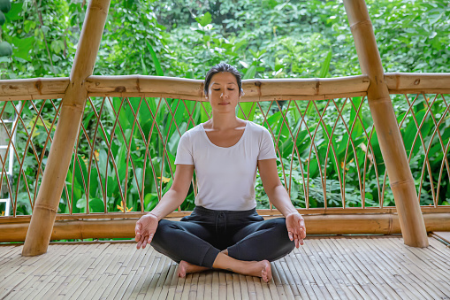 Close-up shot of Southeast Asian Woman meditating with closed eyes, sitting on the floor in a lotus position at a balcony of a sustainable bamboo resort. She crossed her legs and her hands in gyan mudra position.