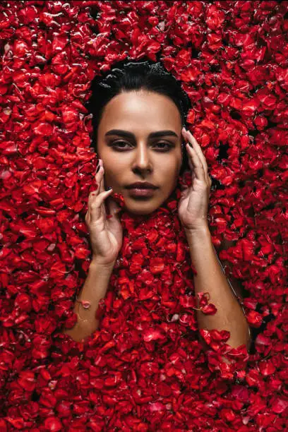 Close-up, top view, beauty portrait, face of a young woman in a bath with water and floating red rose petals and rosebuds. The concept of skin rejuvenation, spa treatments.