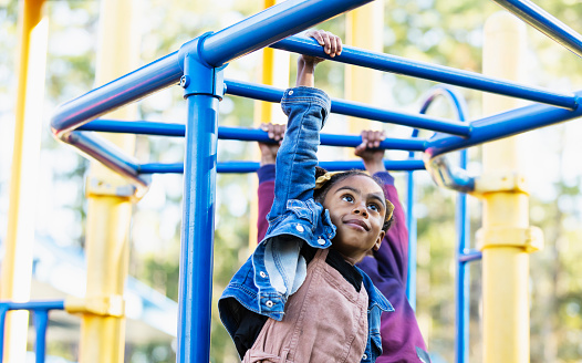 A 5 year old girl playing on a playground, hanging on monkey bars. She is mixed race Hispanic, African-American and Native American.