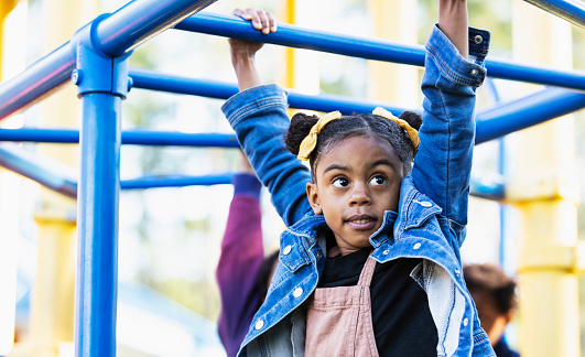 A 5 year old girl playing on a playground, hanging on monkey bars. She is mixed race Hispanic, African-American and Native American.