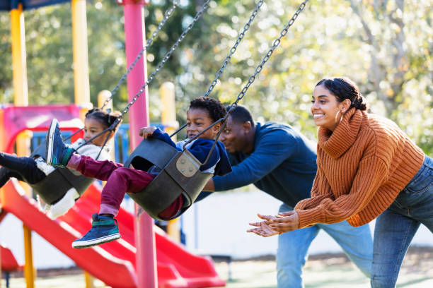 Young family with twins on playground swings A young family playing on a playground. The children, 3 year old twins, are sitting on swings with the parents standing behind them. The focus is the mother and son in the foreground. The father and daughter are out of focus in the background. The children are mixed race Hispanic, African-American and Native American. happy indian young family couple stock pictures, royalty-free photos & images