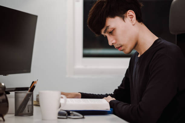 istock Young handsome asian man reading book at work desk late at night, Knowledge and learning concept 1305652165