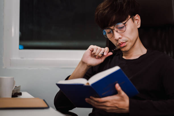 istock Young handsome asian man reading book at work desk late at night, Knowledge and learning concept 1305652693