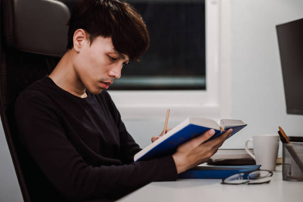 istock Young handsome asian man reading book and writing note at work desk late at night, Knowledge and learning concept 1305652705