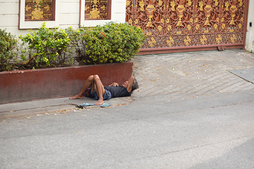 Homeless thai man is laying in street of Bangkok Ladprao in front of a gate to a ground and courtyard. Man is laying close to plants and sewer. He is barefoot,