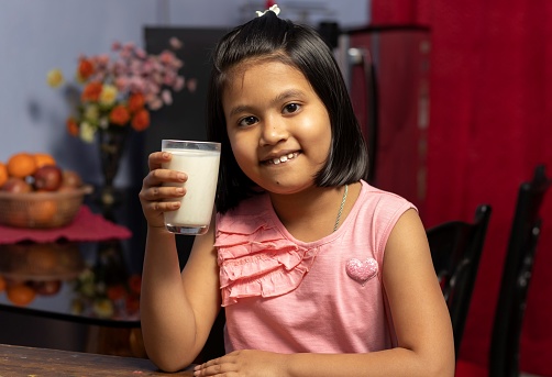 Head shot of a cute little Indian / asian girl holding a glass full of milk with smiling face - healthy eating concept
