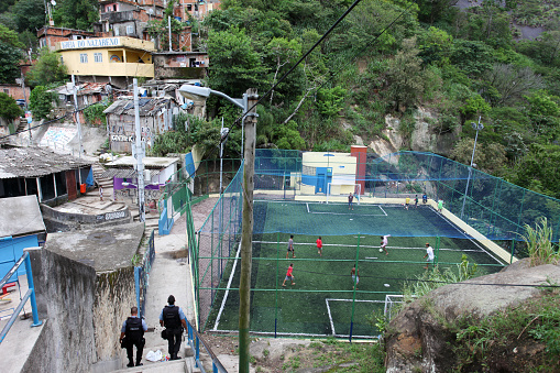 Brazil, Rio de Janeiro - March 4th, 2021: Boys playing soccer at the top of Favela Santa Marta, in Botafogo district. Two policemen go downstairs (left). The Nazareno´s church is located in a yellow house (top left).