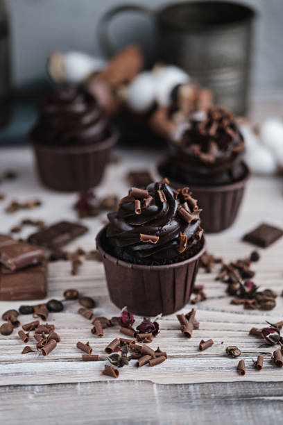 istock Several muffins or cupcakes with chocolate shaped cream at white table. Festive candle burns on a chocolate cake. There are chocolate chips scattered nearby. 1305664549