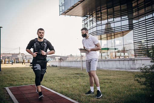 Two people, male personal trainer using digital tablet while his male client is exercising in electrical muscular stimulation suit, in outdoors gym.