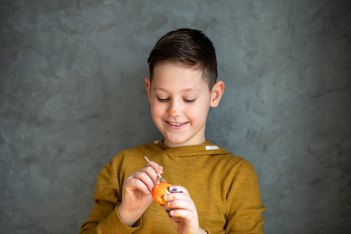 Lovely smiling boy is standing in front of the gray backroad and paints on Easter egg using a paintbrush and orange tempera color. He loves Easter because he has time to be creative and enjoy joint activities with his family.
