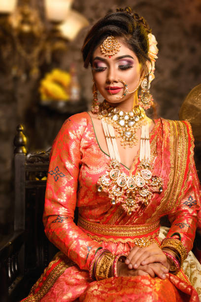 magnificent young indian bride in luxurious bridal costume with makeup and heavy jewellery is sitting in a chair in with classic vintage interior in studio lighting. wedding fashion. - maquiagem ceremonial imagens e fotografias de stock