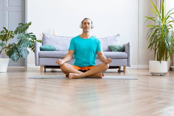 photo d’un homme d’âge moyen pratiquant le yoga à la maison. il est assis dans la position lotus avec un casque et écoute de la musique de méditation - lotus position audio photos et images de collection