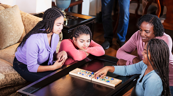 An African-American family with three teenage daughters spending time together at home. It is game night, and they are sitting around the living room coffee table playing mancala. Mother is in her 50s, and the girls are 13, 14 and 16 years old.