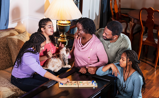 An African-American family with three teenage daughters spending time together at home. It is game night, and they are sitting around the living room coffee table playing mancala. The parents are in their 50s, and the girls are 13, 14 and 16 years old. The family dog, a shih tzu, is hanging out with them.