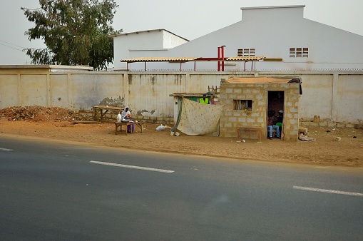 Dakar, Senegal - May 21,2012 : A lone woman sits at a table doing a woman task, while one man stands behind a sheet urinating and two other men sit in the darkness of their home, all against a wall topped with spikes to deter people from climbing over, all surrounded by the ubiquitous trash