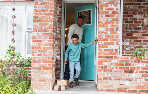 A 5 year old mixed race boy helps his father retrieve packages delivered to their home and left on the front doorstep. He has stepped out onto the porch, with his father standing behind him in the doorway. He is looking down at two boxes. The man is African-American, in his 30s, and his son is African-American and Caucasian.