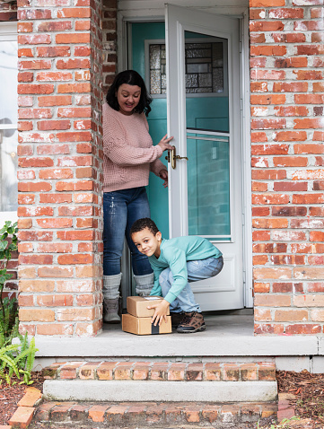 A 5 year old mixed race boy helps his mother retrieve packages delivered to their home and left on the front doorstep. He has stepped out onto the porch, with his mother standing behind him in the doorway. He is bending down and picking up the two boxes while looking at the camera. The woman is Caucasian, in her 40s, and her son is African-American and Caucasian.
