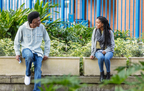 Young man and woman, social distance, holding face masks A young man and woman sitting outdoors, six apart, looking at each other. They are social distancing, holding protective face masks in their hands, trying to prevent the spread of coronavirus during the COVID-19 pandemic. sitting on bench stock pictures, royalty-free photos & images