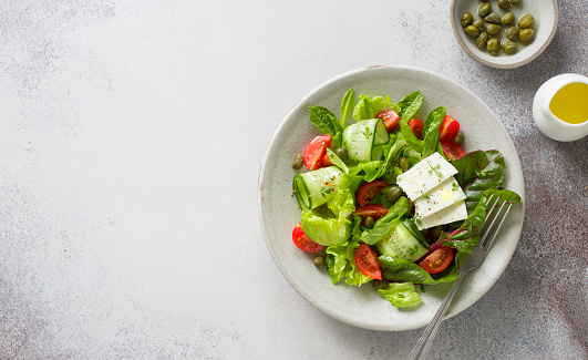 Fresh salad with tomatoes, cucumbers, capers, feta and mixed salad leaves on clay plate  on gray stone background. Flat lay. Copy space