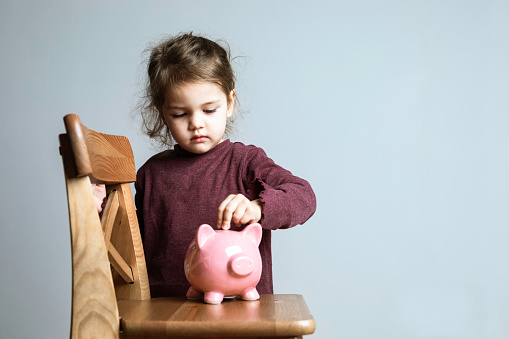 Toddler with is putting coins into her piggy bank in front of a blank gray wall.