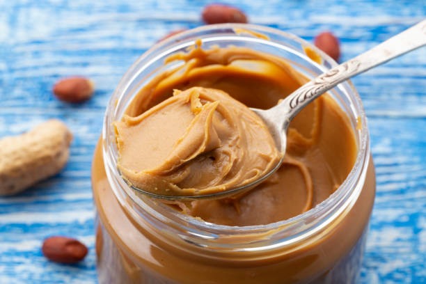 Peanut butter in an open jar and peanuts in the skin are scattered on the blue table. Space for text. stock photo