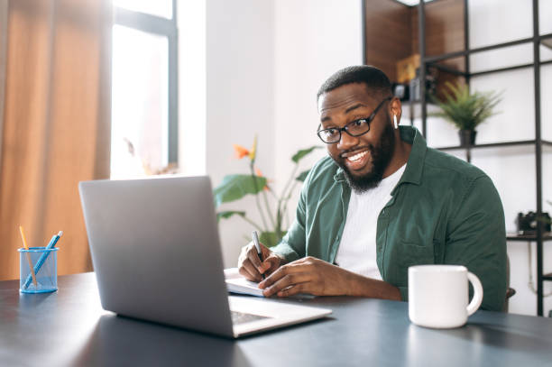 istock Distance learning at home. African American guy student or freelancer uses a laptop for learning, watches a webinar, makes notes in a notebook 1305701571