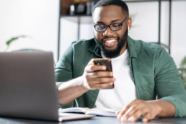 istock Chatting online. Joyful African American guy, sits at his desk, using a mobile phone while working, browsing the internet, answering an email, chatting with friend or family 1305701575