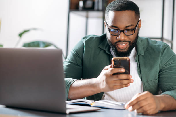 istock Busy confident African American guy, sits at his desk, using a mobile phone while working, browsing the internet, answering an email, looking for ideas for a project 1305701580