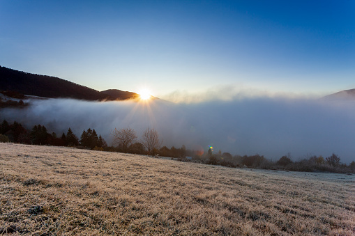 Sun rises from behind mountain top, fog moves and rises up at dawn on summer morning in mountains. Morning fog dissipates in mountains Carpathians. Nature landscape, natural scenery, rural background.